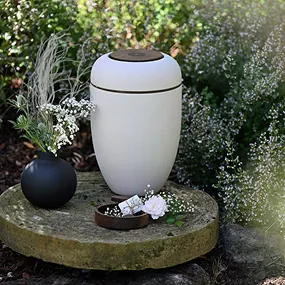 Weiße Urne für Feuerbestattung auf Steinplatte im Garten, daneben schwarze Vase mit weißen Blumen